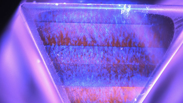 Figure 3. DiamondView image of one of the faces of the stone showing luminescence features consistent with a CVD synthetic diamond. Coarse bands of variable intensity orange emission from nitrogen vacancy centers are visible, along with growth striations and “plumes” of blue emission from dislocations within the bands. Image by Sema Firat; field of view 9.0 mm. Figure 3. DiamondView image of one of the faces of the stone showing luminescence features consistent with a CVD synthetic diamond. Coarse bands of variable intensity orange emission from nitrogen vacancy centers are visible, along with growth striations and “plumes” of blue emission from dislocations within the bands. Image by Sema Firat; field of view 9.0 mm.