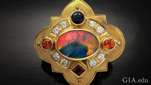 An orange, blue and green stone is the center piece of this shield-like brooch. The center stone is framed by diamonds, orange, blue and red gems. 