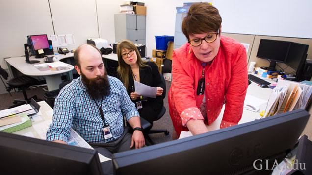 Three jewelry manufacturing arts team members (2 seated, one standing) in front of two large computer monitors.
