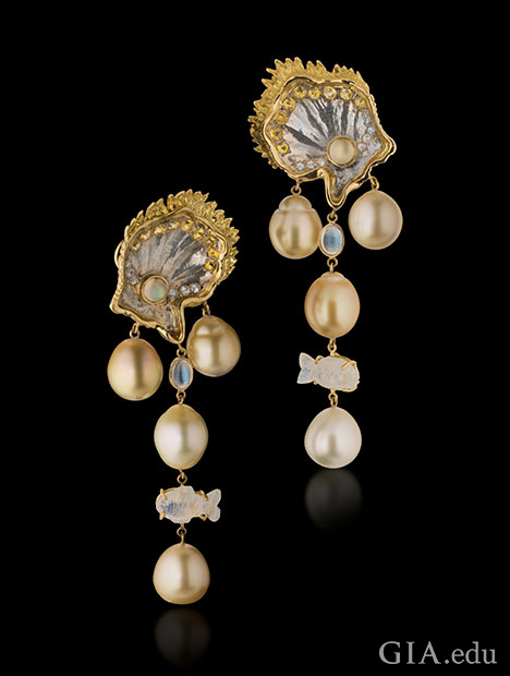South Sea pearls dangle from clam-shapes made from mother of pearl and accented with white and yellow sapphires and 18K yellow gold. Moonstone accents, including one fish-shaped item, also dangle from the top of the earrings.
