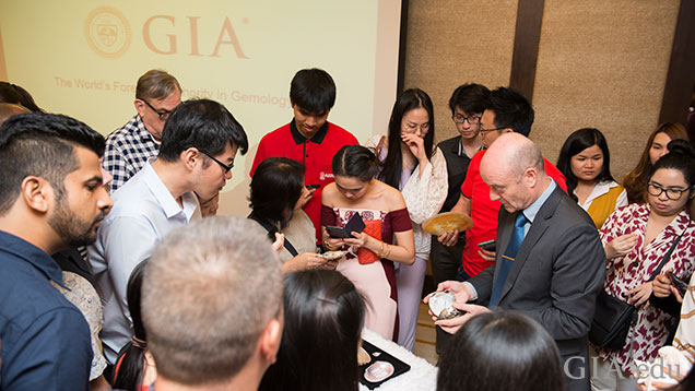 More than a dozen people surround a table of pearl and mollusk samples. More than a dozen people surround a table of pearl and mollusk samples.