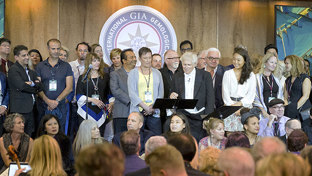 A diverse group of people stands together on stage, with a backdrop featuring the International GIA Gemological logo.