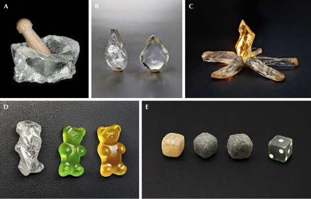 Figure 7. Rough diamonds resembling objects: mortar, 1.95 ct, with a pestle carved from a toothpick (A); raindrops, 3.23 ct each (B); campfire flame, 2.70 ct, backlit using orange light and placed with raw baguettes from figure 3 as firewood (C); gummy bear, 9.18 ct (D; colored gummy bears for comparison created using Adobe Photoshop Generative AI); fibrous diamond cuboids resembling dice, 22.45, 22.47, and 23.64 ct, respectively, shown with a superimposed 10 mm die (E). Photos by Evan M. Smith. Figure 7. Rough diamonds resembling objects: mortar, 1.95 ct, with a pestle carved from a toothpick (A); raindrops, 3.23 ct each (B); campfire flame, 2.70 ct, backlit using orange light and placed with raw baguettes from figure 3 as firewood (C); gummy bear, 9.18 ct (D; colored gummy bears for comparison created using Adobe Photoshop Generative AI); fibrous diamond cuboids resembling dice, 22.45, 22.47, and 23.64 ct, respectively, shown with a superimposed 10 mm die (E). Photos by Evan M. Smith.