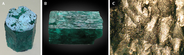 Figure 5. Sample 2 (18.0 mm in length), a columnar emerald that has been heavily corroded by natural etching. A: The basal plane shows irregularly shaped openings to larger cavities below this face. B: The m prism planes show etch structures, especially irregularly oriented grooves between hillocks. C: Details of the etch structures with grooves and hillocks, seen on the m prism face in image B, which are covered by micro-steps as seen in reflected light. Photos by K. Schmetzer; field of view 3.5 mm (C). Figure 5. Sample 2 (18.0 mm in length), a columnar emerald that has been heavily corroded by natural etching. A: The basal plane shows irregularly shaped openings to larger cavities below this face. B: The m prism planes show etch structures, especially irregularly oriented grooves between hillocks. C: Details of the etch structures with grooves and hillocks, seen on the m prism face in image B, which are covered by micro-steps as seen in reflected light. Photos by K. Schmetzer; field of view 3.5 mm (C).