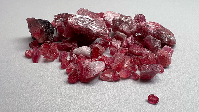 Figure 1. A faceted pezzottaite, measuring approximately 6 × 4 mm, with a group of rough (largest piece ~25 mm in diameter) exhibiting purplish pink to red color from the Vakinankaratra region of Madagascar. Photo by Nathan Renfro; courtesy of Lamine Kakro and Yianni Melas.
