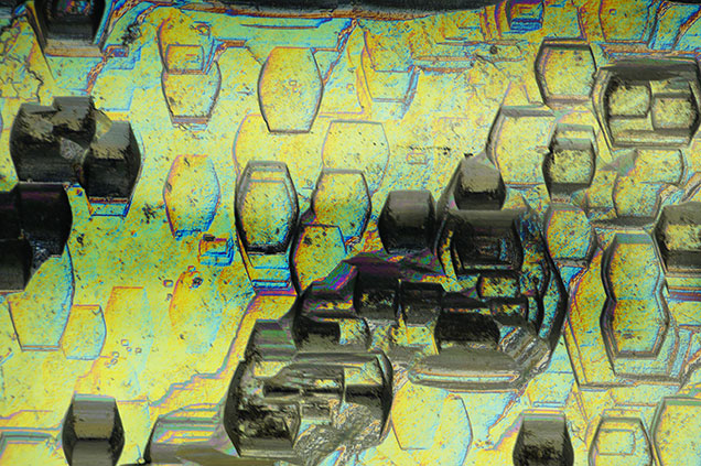 Figure 2. Heliodor viewed with differential interference contrast filters. Compressed hexagonal etch features exist on every prismatic face and run the length of the crystal. Photomicrograph by Rhiana Henry; field of view 2.88 mm. Figure 2. Heliodor viewed with differential interference contrast filters. Compressed hexagonal etch features exist on every prismatic face and run the length of the crystal. Photomicrograph by Rhiana Henry; field of view 2.88 mm.