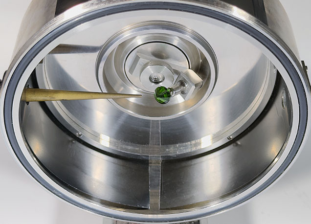 A close-up view of a metallic chamber with a green object at the center, featuring a tool pointing towards it.
